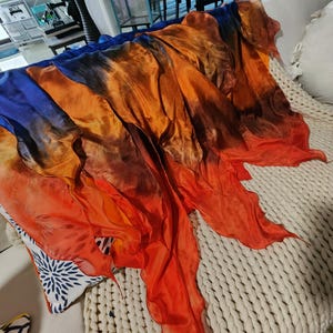 Puede incluir: Una bufanda vibrante de seda en tonos azules, naranjas y rojos. La bufanda está drapeada sobre una superficie texturizada de color crema. Los colores se mezclan, creando un efecto degradado.