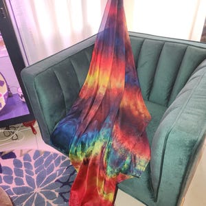 May include: A long, rectangular scarf featuring a vibrant, multi-colored tie-dye pattern. The fabric displays a blend of deep blues, bright reds, yellows, and greens, creating a swirling, nebula-like effect across the entire surface of the material.