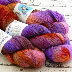 Pode incluir: Um novelo de fio tingido à mão em tons de roxo, rosa e laranja. O fio está etiquetado com "Handgefärbte Wolle" e "Handarbeite Wolle".