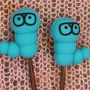 Könnte beinhalten: Zwei blaue, wurmförmige Maschenmarkierer aus Kunststoff mit schwarzen Brillen und lächelnden Gesichtern. Die Markierer sind an bunten Stricknadeln befestigt.