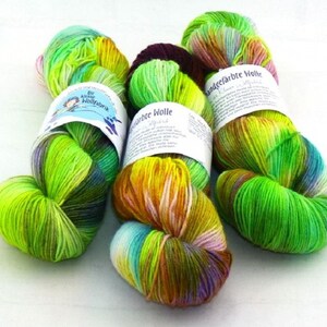 May include: Three skeins of hand-dyed yarn in shades of green, yellow, orange, and purple. The yarn is labelled "Handgefarbte Wolle" and "Die Kleine Wollfabrik".