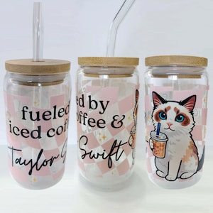May include: Three glass tumblers with bamboo lids and clear glass straws. The tumblers feature pink and white checkered patterns and text that reads "fueled iced coffee Taylor G", "fueled by coffee & Swift", and a cat holding a drink.