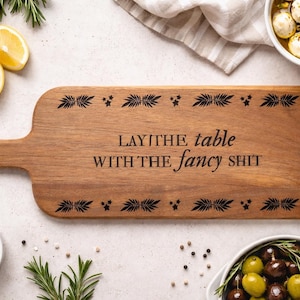 May include: Wooden serving board with a handle, engraved with the phrase "LAY THE table WITH THE fancy SHIT". The board is surrounded by ingredients like lemons, olives, and herbs, suggesting a culinary theme.