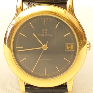 May include: A gold Zenith automatic wristwatch with a black face and a date window at the 3 o'clock position. The watch features gold hands and markers, a black leather strap, and the word "ZENITH" is displayed on the dial.