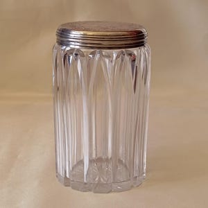 Può includere: Un barattolo di vetro trasparente, cilindrico, con dettagli verticali a coste. Ha un coperchio in metallo color argento. La base del barattolo presenta un disegno a stella. Lo sfondo è beige chiaro.