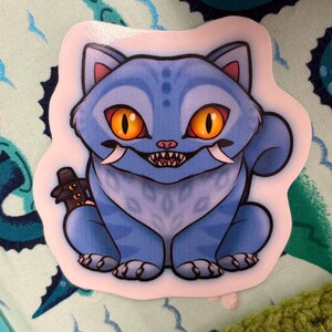 以下が含まれることがあります： オレンジ色の目、牙、牙を持つ漫画の猫が描かれた青いステッカー。猫は小さな暗いオブジェクトを持っています。ステッカーは白い縁取りがあり、模様のある背景に配置されています。