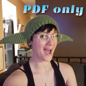 May include: A person wearing a green crocheted hat with large ears, resembling a character from Star Wars. The hat is green and has a crocheted texture. The person is wearing glasses and has a happy expression.