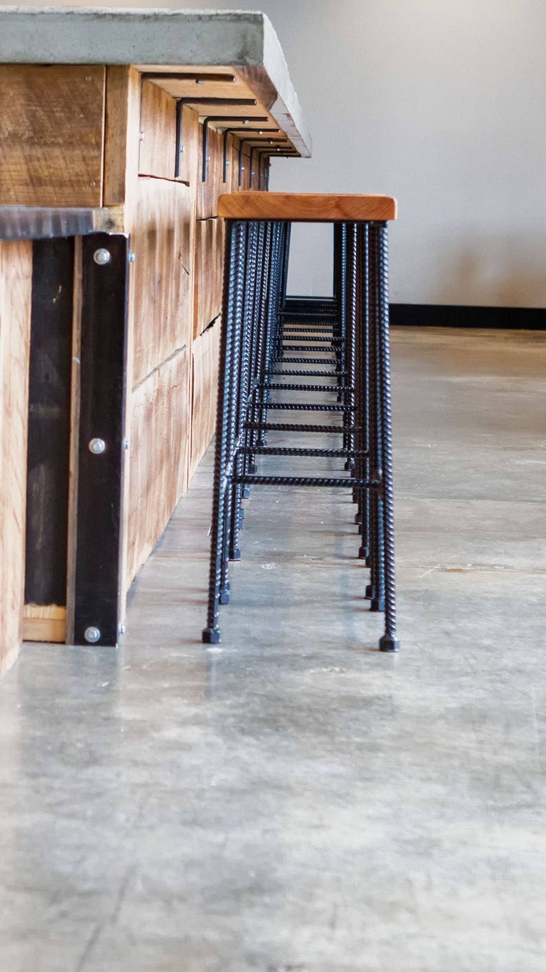 May include: A row of black metal bar stools with wooden seats line up against a wooden counter with a concrete top.