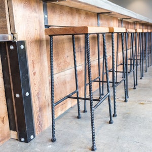 May include: A row of black metal bar stools with wooden seats lined up against a wooden wall. The stools have a simple, industrial design.
