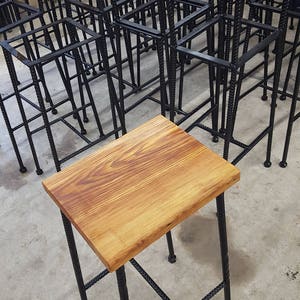 May include: A wooden stool with a natural wood top and black metal legs. The legs are made of twisted metal and have a geometric design.