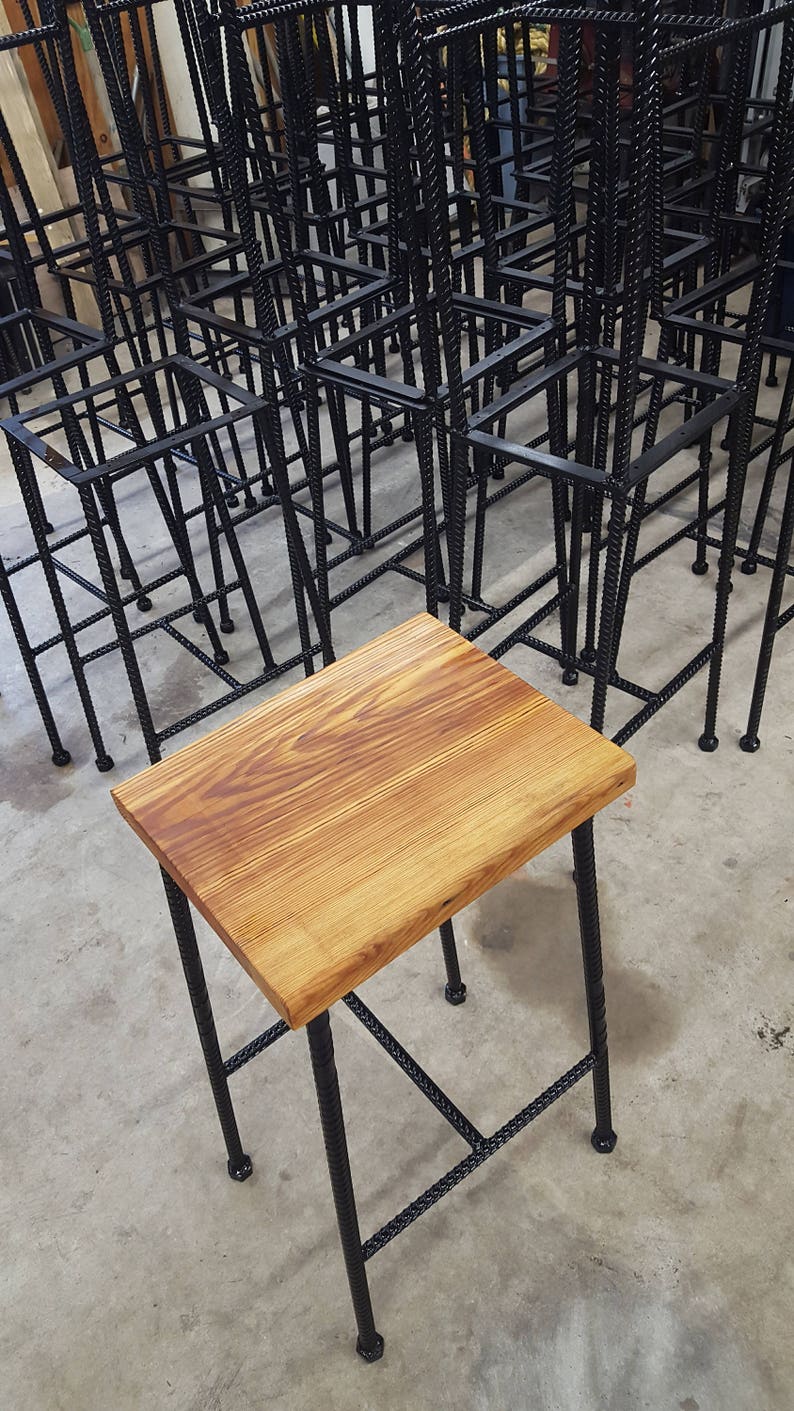May include: A wooden stool with a natural wood top and black metal legs. The legs are made of twisted metal and have a geometric design.