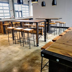 May include: A long wooden bar with black metal supports and stools. The bar is made of reclaimed wood and has a rustic look. The stools are black metal with wooden seats.