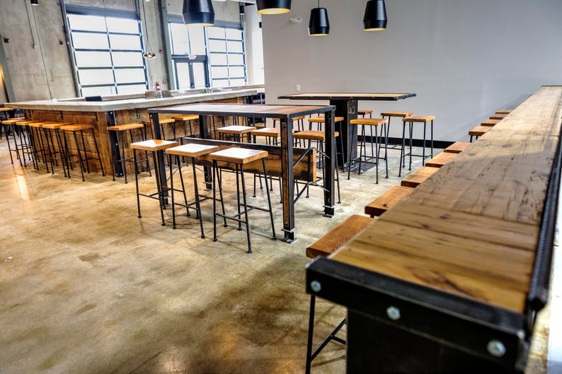 May include: A long wooden bar with black metal supports and stools. The bar is made of reclaimed wood and has a rustic look. The stools are black metal with wooden seats.