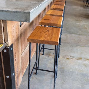 May include: A row of wooden bar stools with black metal frames. The stools are lined up in front of a concrete countertop.
