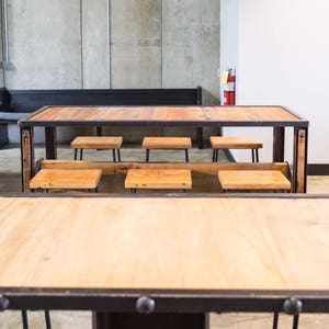 May include: A long wooden table with black metal legs and a black metal frame. The table is surrounded by six wooden stools with black metal legs. The table and stools are in a room with concrete walls and black pendant lights.