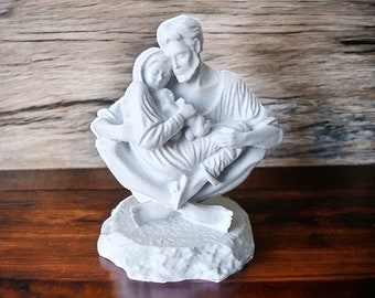 May include: A white ceramic sculpture of a family, depicting a man, woman, and child. The family is seated together, with the man holding the child. The sculpture is on a white base.