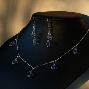 K&ouml;nnte beinhalten: Eine silberne Halskette mit einer zarten Kette und mehreren tropfenf&ouml;rmigen Kristallanh&auml;ngern. Passende silberne Ohrringe mit Kristalltropfen h&auml;ngen an den Haken.