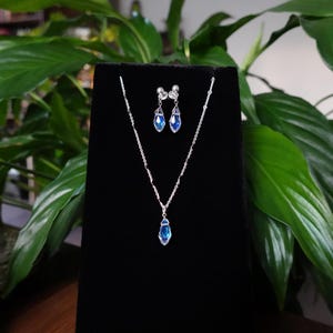 May include: A silver necklace and earrings set with blue teardrop-shaped crystals. The jewellery is displayed on a black velvet background.