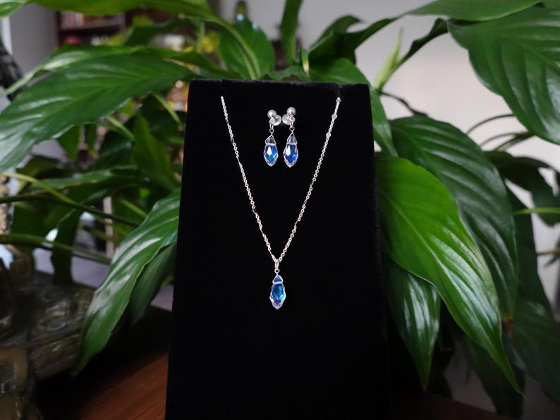 May include: A silver necklace and earrings set with blue teardrop-shaped crystals. The jewellery is displayed on a black velvet background.