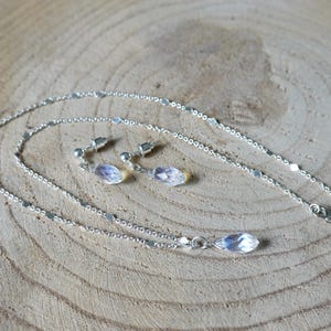 May include: A silver chain necklace with a teardrop-shaped crystal pendant and matching earrings. The necklace and earrings are on a wooden surface.