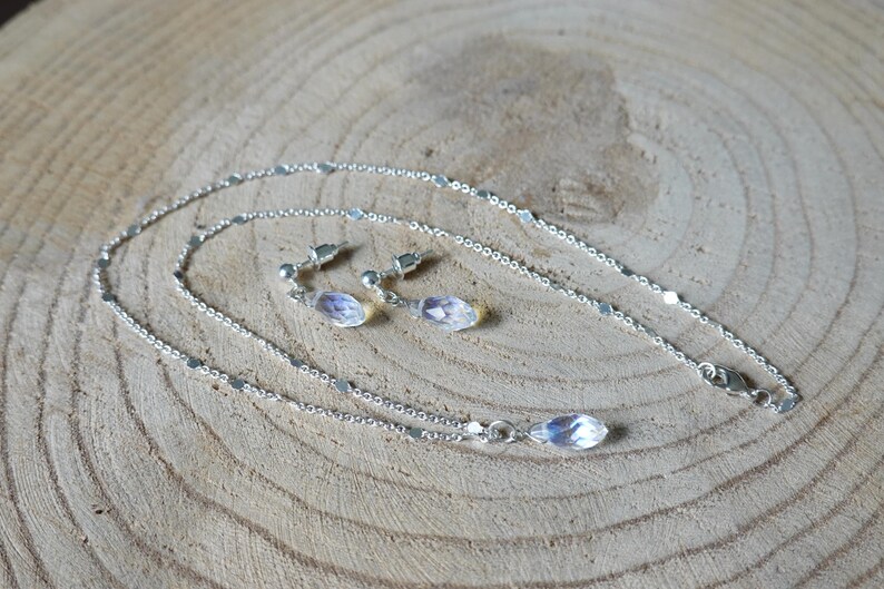 May include: A silver chain necklace with a teardrop-shaped crystal pendant and matching earrings. The necklace and earrings are on a wooden surface.