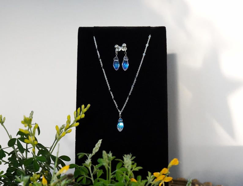 May include: A silver necklace and earrings set with blue teardrop-shaped crystals on a black velvet display stand.