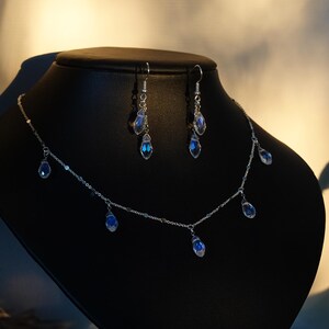 K&ouml;nnte beinhalten: Eine silberne Halskette mit einer zarten Kette und f&uuml;nf baumelnden blauen Tropfenkristallen. Passende Ohrringe mit zwei Tropfenkristallen h&auml;ngen an silbernen Ohrsteckern.