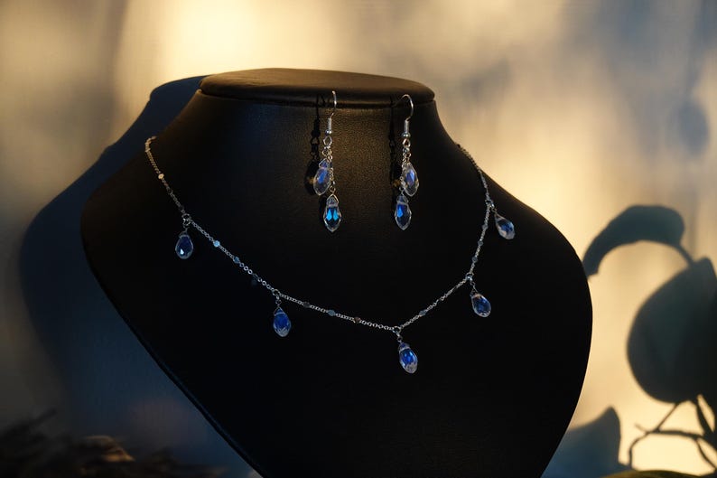K&ouml;nnte beinhalten: Eine silberne Halskette mit einer zarten Kette und f&uuml;nf baumelnden blauen Tropfenkristallen. Passende Ohrringe mit zwei Tropfenkristallen h&auml;ngen an silbernen Ohrsteckern.
