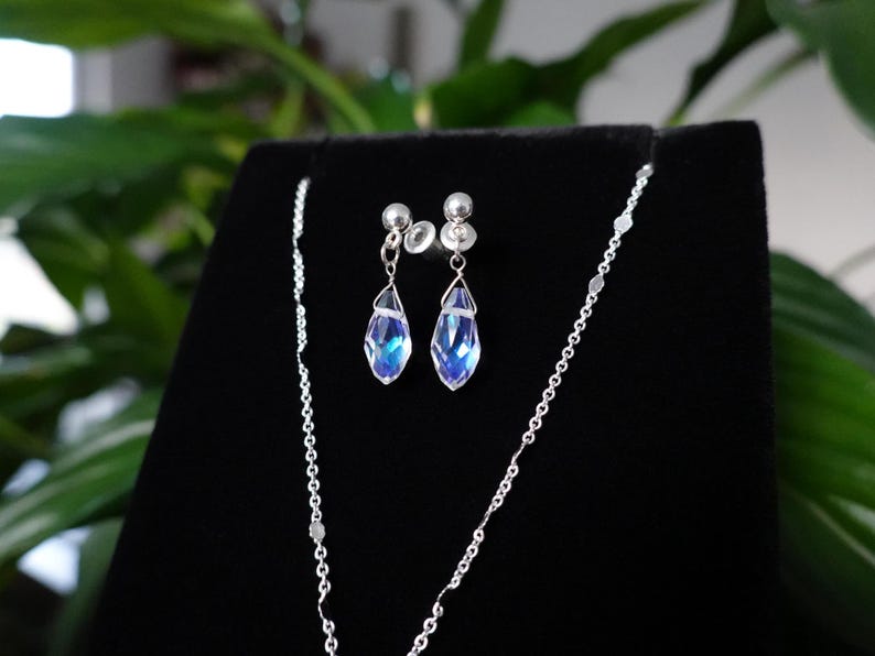May include: A silver necklace and earrings set with teardrop-shaped blue crystals. The necklace is a delicate chain with a small pendant.