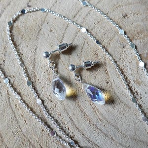 May include: A silver chain necklace with a delicate chain and small silver discs. The necklace is paired with a set of silver earrings with teardrop-shaped clear crystals.