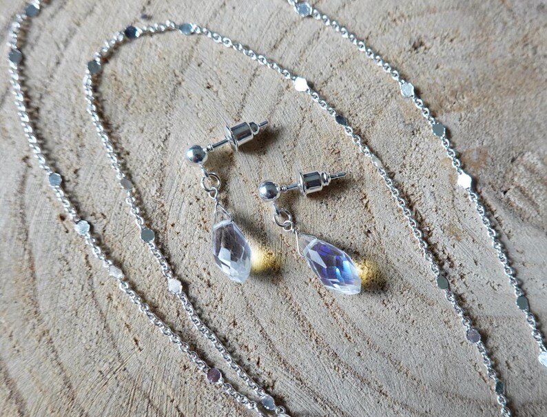 May include: A silver chain necklace with a delicate chain and small silver discs. The necklace is paired with a set of silver earrings with teardrop-shaped clear crystals.