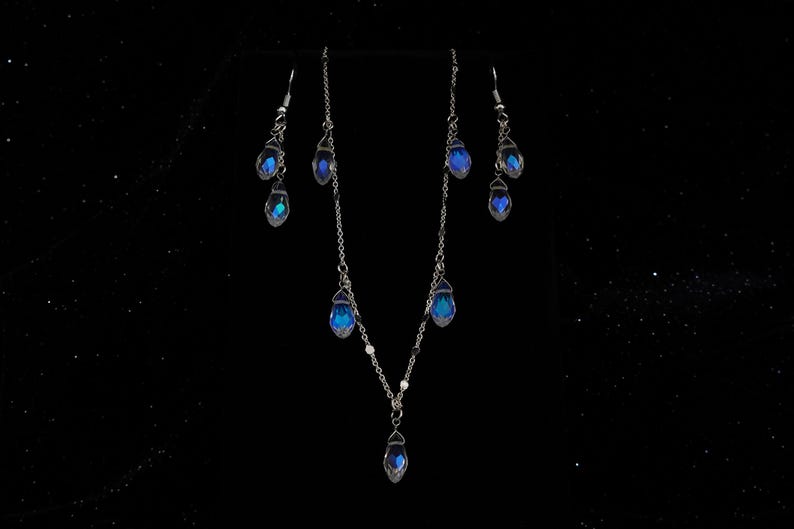 K&ouml;nnte beinhalten: Ein silbernes Halsketten- und Ohrring-Set mit blauen, tropfenf&ouml;rmigen Kristallen. Die Halskette hat einen einzelnen Kristall-Anh&auml;nger und die Ohrringe haben jeweils zwei Kristalle.