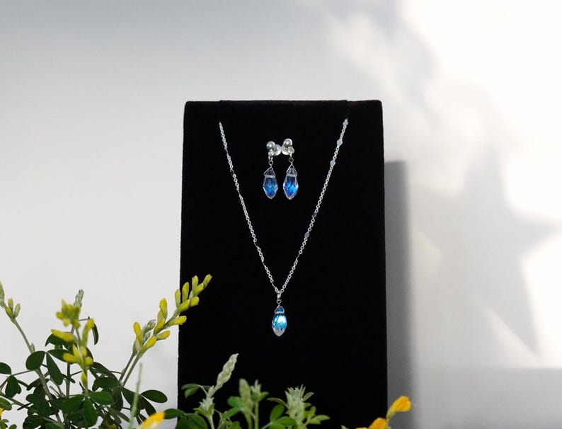 May include: A silver necklace and earrings set with blue teardrop-shaped crystals on a black velvet display stand.