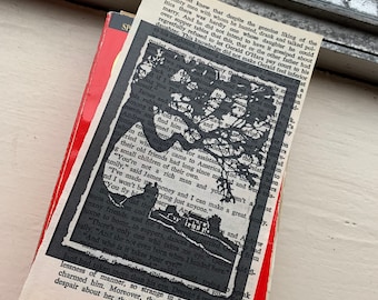 May include: A vintage book with a red cover and a black and white cutout image of a tree and house on top. The cutout is placed over the pages of the book, with text visible.