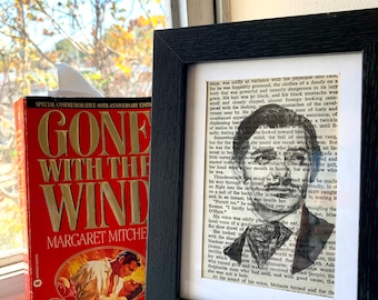 May include: A framed black and white portrait of a man, set against a backdrop of printed text. Beside it, a red book with gold lettering, titled "Gone With The Wind" by Margaret Mitchell, is propped up.