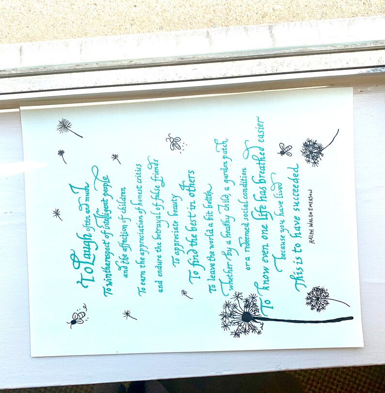 Op de afbeelding: Een wit ingelijst print met een turquoise citaat over leven en lachen. Het citaat bevat de tekst "To laugh often and much" en "This is to have succeeded". Het citaat is omringd door zwart-wit illustraties van paardenbloemen.