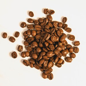 Peut inclure: Vue de dessus d'un tas de grains de caf&eacute; torr&eacute;fi&eacute;s. Les grains sont d'un brun fonc&eacute; riche, certains pr&eacute;sentant des reflets plus clairs. Ils sont dispos&eacute;s sur une surface blanche, cr&eacute;ant une esth&eacute;tique naturelle et organique.
