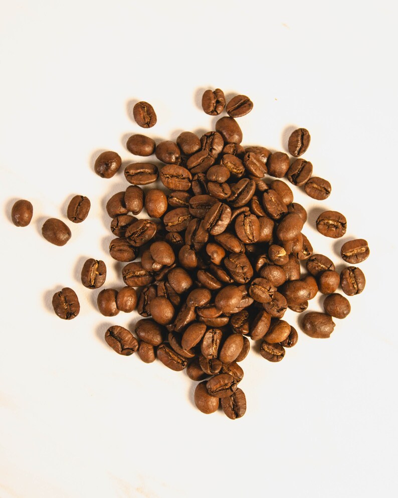 Peut inclure: Vue de dessus d'un tas de grains de caf&eacute; torr&eacute;fi&eacute;s. Les grains sont d'un brun fonc&eacute; riche, certains pr&eacute;sentant des reflets plus clairs. Ils sont dispos&eacute;s sur une surface blanche, cr&eacute;ant une esth&eacute;tique naturelle et organique.
