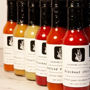 May include: A row of glass bottles filled with various colored hot sauces. Each bottle has a black cap and a white label with the text "COLDFIRE HOT SAUCE" and a flame logo. The sauces range in color from orange to red.