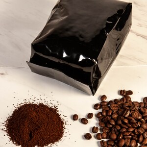 Peut inclure: Un sac de caf&eacute; noir brillant est plac&eacute; au-dessus d'un tas de caf&eacute; moulu et d'un groupe de grains de caf&eacute; entiers. Le caf&eacute; moulu est brun fonc&eacute;, et les grains sont d'un brun torr&eacute;fi&eacute; riche. Le fond est une surface en marbre blanc.