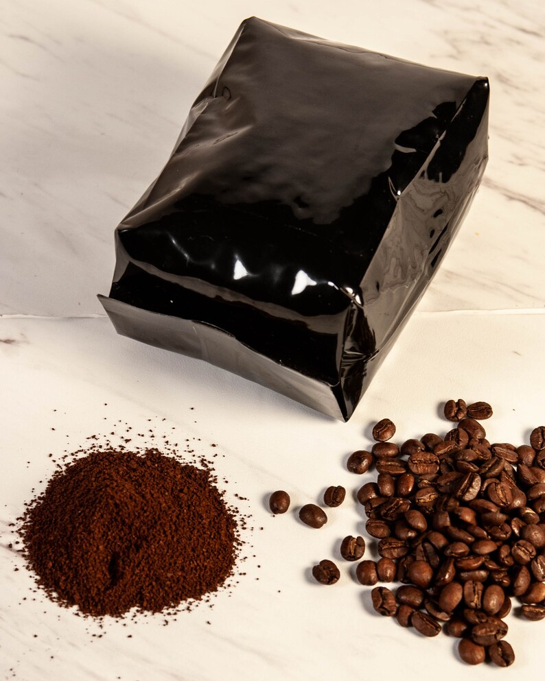 Peut inclure: Un sac de caf&eacute; noir brillant est plac&eacute; au-dessus d'un tas de caf&eacute; moulu et d'un groupe de grains de caf&eacute; entiers. Le caf&eacute; moulu est brun fonc&eacute;, et les grains sont d'un brun torr&eacute;fi&eacute; riche. Le fond est une surface en marbre blanc.