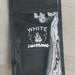 May include: A black, sealed pouch with a white label featuring a skull design and the words "WHITE LIGHTNING" in bold, stylized font. The pouch has a shiny, reflective surface and is lying on a light-colored wooden surface.