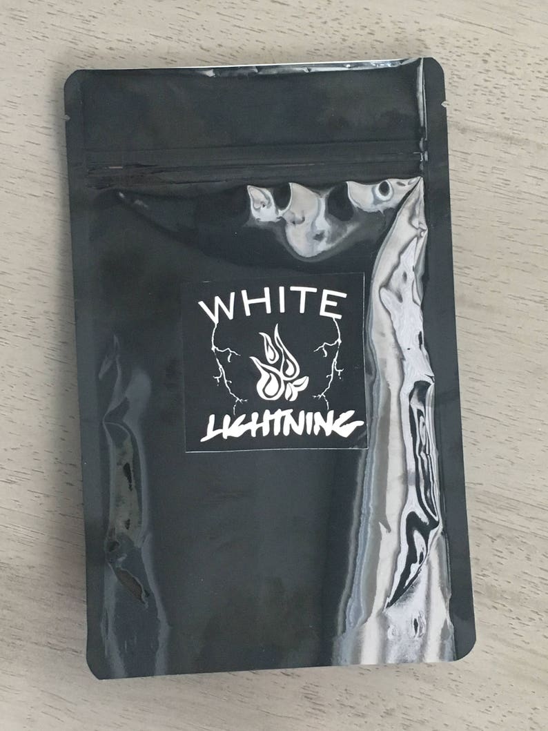 May include: A black, sealed pouch with a white label featuring a skull design and the words "WHITE LIGHTNING" in bold, stylized font. The pouch has a shiny, reflective surface and is lying on a light-colored wooden surface.