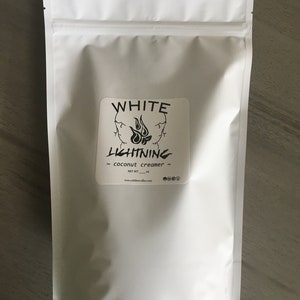 May include: A white, resealable pouch of coconut creamer. The label features the text "WHITE LIGHTNING" above a flame graphic. The words "coconut creamer" are below the graphic. The net weight is listed in ounces. The website address is also printed on the label.