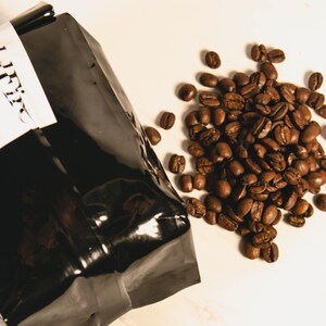 Peut inclure: Un sac de caf&eacute; noir avec une &eacute;tiquette blanche et un tas de grains de caf&eacute; torr&eacute;fi&eacute;s. Les grains sont d'une riche couleur brune et sont dispers&eacute;s sur une surface blanche. Le sac est partiellement ouvert, sugg&eacute;rant qu'il contient des grains de caf&eacute;.