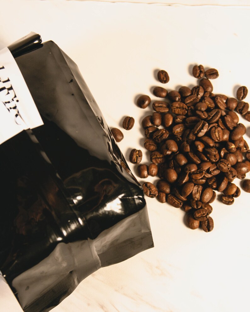 Peut inclure: Un sac de caf&eacute; noir avec une &eacute;tiquette blanche et un tas de grains de caf&eacute; torr&eacute;fi&eacute;s. Les grains sont d'une riche couleur brune et sont dispers&eacute;s sur une surface blanche. Le sac est partiellement ouvert, sugg&eacute;rant qu'il contient des grains de caf&eacute;.
