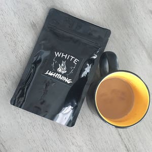 May include: A black pouch with the text "WHITE LIGHTNING" and a flame logo next to a black mug with a yellow interior filled with a light brown liquid. The items are on a light gray wooden surface.
