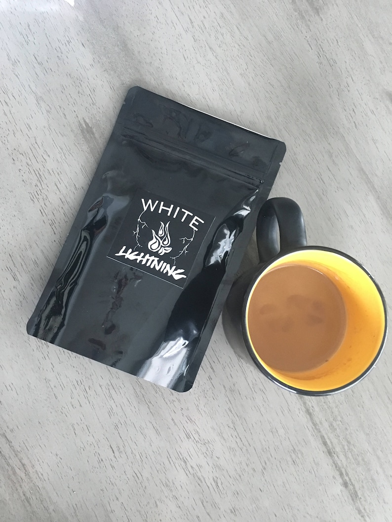 May include: A black pouch with the text "WHITE LIGHTNING" and a flame logo next to a black mug with a yellow interior filled with a light brown liquid. The items are on a light gray wooden surface.