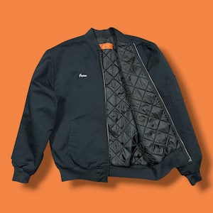 May include: A black bomber jacket featuring a ribbed collar, cuffs, and hem. The front displays the name Juan embroidered in white script. The interior is lined with black quilted fabric, and a bright orange label is visible at the neck.