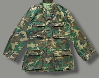 Puede incluir: Chaqueta militar de camuflaje en tonos verdes, marrones y beiges. La chaqueta tiene múltiples bolsillos delanteros y las letras "USMC" impresas en el pecho. La chaqueta tiene cierre de botones y mangas largas.